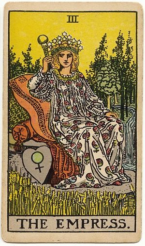 The Empress: A crowned woman sits on cushions in a field of ripe wheat, beside her a heart-shaped shield with the sign of Venus, and behind her a forest and a waterfall.